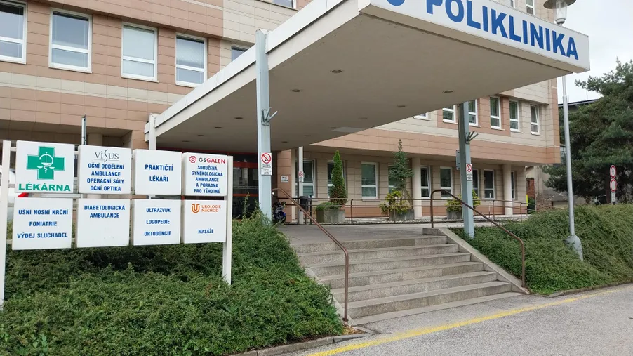 Vstup do budovy polikliniky s nápisem „POLIKLINIKA“, schody a značením zdravotnických služeb včetně lékárny.